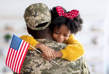 Happy Little Black Daughter Hugging Her Soldier Dad After He Came Back Home From US Army, Smiling Girl Holding American Flag And Embracing Father In Military Uniform, Celebrating Reunion With Daddy