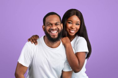 Sevgi ve ilgi. Milenyum siyahi kadının, erkeğini kucaklayan beyaz tişörtlü çiftin portresi mavi stüdyo arka plan duvarında izole bir şekilde kameraya bakıyor ve poz veriyor.