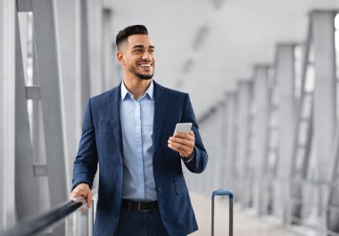 Havalimanı Terminalinde Ellerinde Akıllı Telefonlu Yakışıklı Orta Doğulu, gülümseyen Arap İş Adamı Cep telefonunu tutuyor ve uzaklara bakıyor, Çevrimiçi İletişimin keyfini çıkarıyor, Boşluğu Kopyala