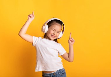 Keyfim yerinde. Sevimli Asyalı çocuk müzik dinliyor, kablosuz kulaklık takıyor ve dans ediyor, sarı duvarın üzerinde izole edilmiş.