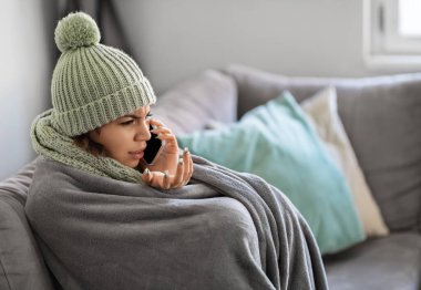 Battaniyeye sarılı bir şekilde koltukta otururken cep telefonuyla konuşan stresli genç kadın, evde donan kadın kiracı, ev sahibine oda sıcaklığının düşük olması, yan görünüşü hakkında şikayette bulundu.