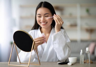 Güzel Asyalı kadın göz serumu uyguluyor ve aynaya gülümsüyor, beyaz bornoz giyiyor, güzellik ürünleriyle dolu bir şişe tutuyor, fotokopi odası. Yaşlanma karşıtı özünü kullanan çekici Koreli kadın.