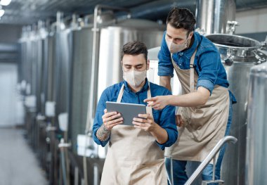 Bira fabrikası çalışanları verileri tablete yükler ve ekipmanları çalıştırırlar. Koruyucu maskeli ve önlüklü gençler COVID-19 salgını sırasında fabrikanın içindeki bira tanklarını tartışıyor ve onunla çalışıyorlar.