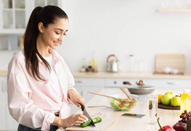 Modern mutfakta lezzetli ve sağlıklı yemekler pişiren mutlu genç bir kadının portresi. Gülümseyen ev kadını sebze salatası hazırlıyor, salatalığı bıçakla doğruyor.