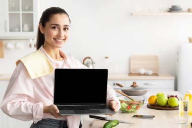 Neşeli genç bayan boş dizüstü bilgisayar pozu gösteriyor. Modern mutfakta duruyor. Gülümseyen Kadın Mutfak Uygulaması Öneriyor ya da Sağlıklı İnternette Tarifleri Olan Web Sitesi. Model