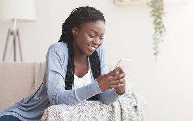 Gülümseyen Afrikalı Amerikalı kadın evinde akıllı telefon kullanıyor. Kanepede dinlenirken mesajlaşıyor ya da sosyal ağlara bakıyor.