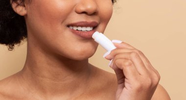 Kıvırcık saçlı bir kadın dudaklarını nemlendirmek için beyaz dudak kremi kullanıyor. Yüzündeki ifade cilt bakımını gösteriyor. Sıcak, nötr bir ortamda, kırpılmış.