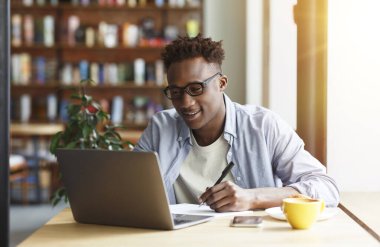 E-öğrenme konsepti. Genç Afro-Amerikan öğrenci laptopuyla kafede kahvaltı sırasında not alıyor.