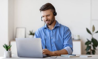 Çevrimiçi kulaklıklı yakışıklı genç adam, dizüstü bilgisayarda Webinar izliyor, uzaktan eğitimin tadını çıkarıyor ve internetten öğreniyor, evde masa başında oturuyor, bilgisayar ekranına bakıyor, boş alana bakıyor.