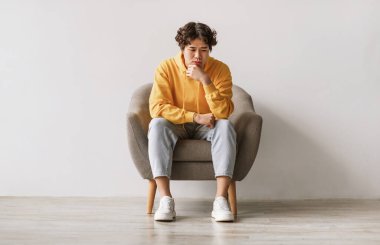 Olumsuz duygular. Asyalı genç bir adam düşünceli bir yüz ifadesiyle koltukta oturuyor, üzgün ya da yorgun hissediyor, problem yaşıyor, depresyon yaşıyor ya da beyaz stüdyo duvarına karşı stres yaşıyor.