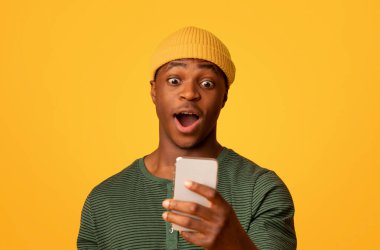 Şok edici bir teklif. Heyecanlı siyah hipster adam akıllı telefon ekranına bakıyor, şaşırmış Afrikalı Amerikalı adam sarı arka planda ağzı açık duruyor, duygusal tepki veriyor, internetteki haberleri kopyalıyor.