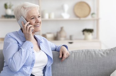 Modern İletişim. Evde cep telefonuyla konuşan gülümseyen yaşlı kadın oturma odasında dinleniyor, boşluğu kopyalıyor.