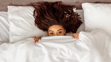 Neşeli Sabah Havası. Güzel Afro-Amerikan kadın portresinin üstünde beyaz çarşafların üzerine uzanmış, yüzü battaniyeyle örtülmüş, dışarı bakıyor, eğleniyor ve saklanıyor.