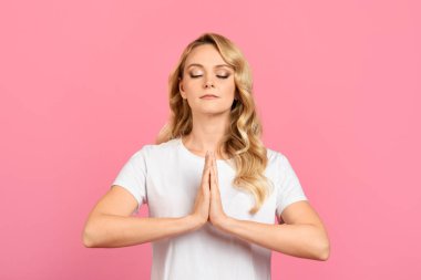 Bir kadın durgun, gözleri kapalı, farkındalık meditasyonu yapıyor. Sarışın ve sade beyaz bir gömlek giyiyor. Sakinleştirici pembe bir arka planı var. Huzur ve sükuneti temsil ediyor..