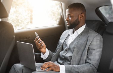 İş seyahatinde bilgisayarlı akıllı telefonu olan yakışıklı Afro-Amerikan iş adamı, fotokopi alanı. Siyah girişimci cep telefonu ekranına bakıyor, dizüstü bilgisayar kullanıyor