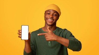 Beyaz ekranlı akıllı telefonu tutan ve işaret eden mutlu siyah hippi adam, Yeni Uygulama veya Mobil Web sitesi öneren neşeli Afrikalı Amerikalı adam, sarı arka planda duruyor,