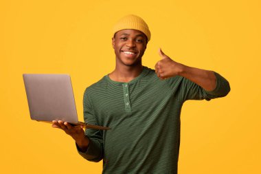 Uzak Çalışma Fırsatları. Elinde dizüstü bilgisayarla gezen neşeli siyah hipster bir adamın portresi. Genç Afrikalı bir adam yeni bir web sitesi tavsiye ediyor. Sarı arka planda duruyor.