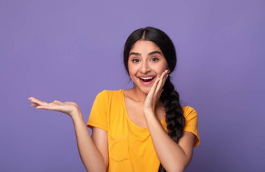 Vay canına, şuna bak. Gülümseyen Hintli genç bir bayanın el ele tutuşması, açık avuçlar, reklam ve boş alan göstermesi ve mor stüdyo duvarında izole edilmiş yanağına dokunması portresi.