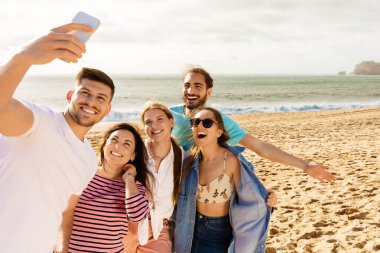 Beş arkadaştan oluşan bir grup kumlu sahilde toplanıyor, gülümsüyor ve arkalarında güneş batarken selfie çekiyorlar, deniz kenarındaki gezileri sırasında neşeli ve canlı bir atmosfer yaratıyorlar..