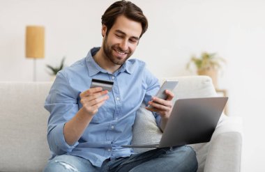 Memnuniyet duyulan adam cep telefonu ve kredi kartı kullanıyor dizüstü bilgisayarla oturuyor ve evde alışveriş yapıyor. Akıllı Telefonlu Müşteri Dükkanları. Mobil Bankacılık Uygulaması ve Ödeme Doğrulama Konsepti