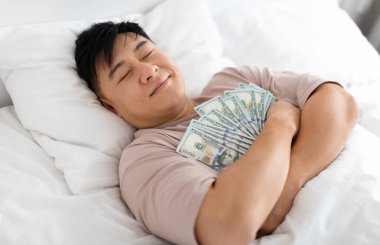 Happy sleeping middle aged chinese man hugging bunch of cash dollars, lying on bed, asian male keeps money safe, top view. Savings, financial literacy, economy concept