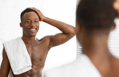 Gömleksiz siyah adam aynaya bakıp saçlarına dokunuyor. Sabah duşundan sonra banyoda dikilirken yansımasına gülümsüyor. Erkek saçı rutini ve güzellik konsepti. Seçici Odaklanma