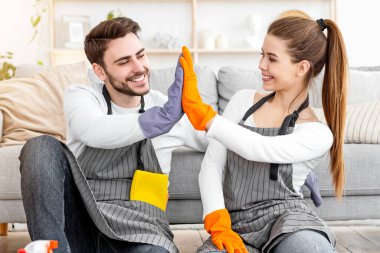 Birlikte eğlenceli temizlik. Önlüklü ve lastik eldivenli gülümseyen adam ve kadın oturma odasının içinde temizlik malzemeleriyle beşlik çakıyor.