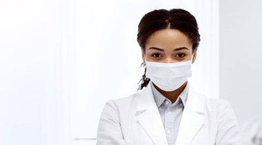 İş yerinde tıbbi maske takan genç siyah kadın stomatolog portresi, profesyonel diş hekimi katlanmış kollarıyla ayakta duruyor ve erkek hastayı kontrol ederken kameraya bakıyor.