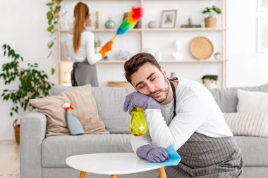 Ev işleri ve yorgunluk. Önlüklü ve eldivenli bir adam dizlerinin üstünde ve masada uyuyor, karısı oturma odasında mobilyaları siliyor, boş yer var.