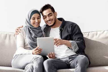 Neşeli müslüman aile yeni dairede koltukta oturuyor, ped kullanıyor, kopyalama alanı kullanıyor. Orta Doğulu mutlu genç adam ve kadın tesettürlü ev için mobilya alıyor taşındıktan sonra eşyaları açıyor.