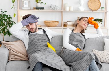 Ev temizliği bitti. Yorgun adam ve kız önlüklü ve lastik eldivenli alnındaki teri silip oturma odasındaki temizlik malzemeleriyle kanepede oturuyor.