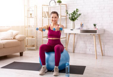 Evde dayanıklılık eğitimi. Milenyum kızı oturma odasında dambıl ve spor topuyla egzersiz yapıyor.