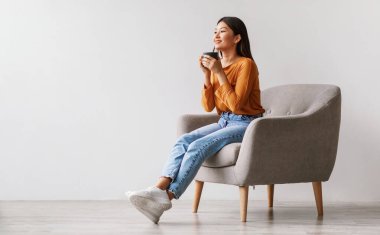 Güzel genç Asyalı kadın kahve ya da çay içiyor, mola veriyor, beyaz stüdyo duvarına yaslanmış koltukta dinleniyor, fotokopi çekiyor. Milenyum dişisi serinliyor ve sıcak içeceğin tadını çıkarıyor.