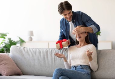 Gülümseyen milenyum Avrupalı adam kadınlara gözlerini kapar, kutuda hediyesini verir, oda içinde gülümseyen eşine sürpriz yapmak için, boş bir alan. Evde parti, doğum günü hediyesi, yıldönümü, Sevgililer günü ve tatil