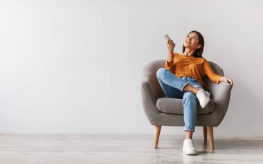 Split system. Happy young Asian woman with remote relaxing under air conditioner, sitting in armchair against white studio wall, mockup. Happy millennial lady using climate control clipart