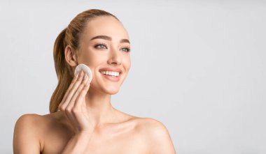 Yüze Güzellik Tedavisi. Pamuk pedi kullanan mutlu genç bayan Gray Stüdyo Arkaplanı 'na yan bakıyor. Panorama, Boş Alan