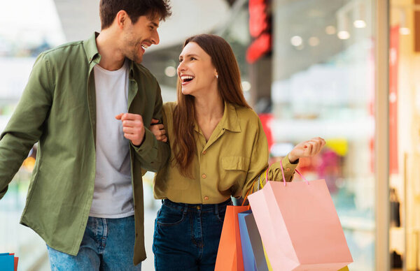Радостная парочка, несущая сумки и улыбающаяся друг другу, проводящая время в торговом центре по выходным. Счастливые покупатели покупают одежду во время сезонных продаж. Концепция торговли и шопоголизма