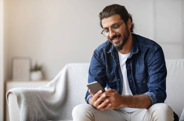 Yakışıklı Hintli genç adam elinde akıllı telefonuyla evde oturuyor, arkadaşlarıyla mesajlaşıyor, koltukta cep telefonuyla dinleniyor, yeni uygulamaları kontrol ediyor, mobil iletişimden keyif alıyor ya da sosyal ağlara takılıyor.