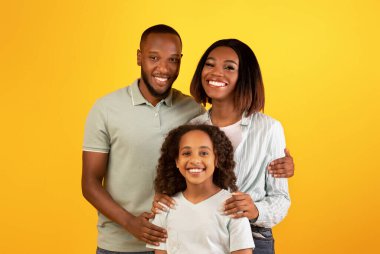 Aile sevgisi kavramı. Sarı arka planda izole edilmiş siyah aile portresi ve kızının kameraya sarılıp gülümsemesi. Annem ve babam stüdyoda çocukla poz veriyorlar.