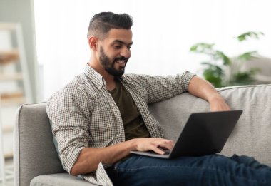 Serbest çalışma, online iş ya da öğrenim kavramı. Kanepede dizüstü bilgisayarıyla oturan neşeli genç bir Arap, evde çalışıyor ya da öğreniyor. Millennial Eastern elemanı Webinar ya da konferans yapıyor.