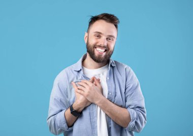 Milenyum adamı elini göğsüne dayayıp mavi stüdyo arka planında olumlu duygular ve minnettarlığını ifade ediyor. Mutlu genç adam avucunu kalbinde tutuyor, umut ve aşkı hissediyor.