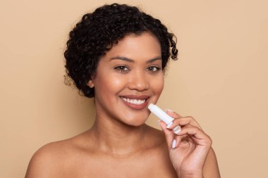 Kıvırcık saçlı bir kadın dudaklarına mutluluk kremi sürüyor. Pürüzsüz bir bej arka planın önünde duruyor, parlak cildini ve neşeli ifadesini sergiliyor..