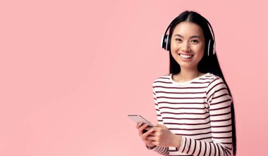 Akıllı telefon ve kablosuz kulaklıkla müzik dinleyen neşeli Asyalı genç kadın pembe stüdyo arka planında otururken mutlu Koreli kadın yeni mobil ses uygulamasının keyfini çıkarıyor.
