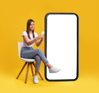 Neşeli gülümseyen kadın boş ekranlı büyük bir cep telefonunun yanında oturuyor, mobil cihaz kullanıyor, turuncu stüdyo arka planındaki yeni uygulamaları kontrol ediyor, web sitesi ya da uygulama tasarımı için makara yapıyor.