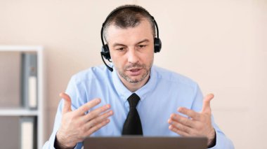 Modern Ofisteki uzak iş arkadaşlarıyla çevrimiçi toplantı sırasında bilgisayarın başında oturup video görüşmesi yapan kulaklıklı bir iş adamı.