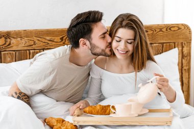 Yatakta kahvaltı. Koca karısını yanağından öpüyor, mutlu eş kahve koyuyor.
