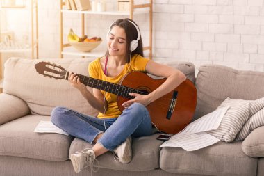 Evde kalıp boş vakit geçirin. Oturma odasındaki kanepede gitar çalan kulaklıklı, gülümseyen kız.