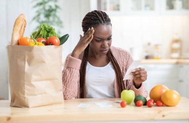 Market masrafları. Mutfakta masada oturan endişeli siyahi kadın, yemek alışverişinden sonra Bill 'i kontrol eden Afrikalı Amerikalı kadın, yüksek fiyatlar, finansal kriz, yoksulluk ve bedava alan konusunda endişeli.