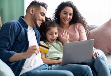 Bilgisayarı ve kredi kartı olan neşeli Arap ailesi evdeki koltukta dinlenirken internetten alışveriş yapıyor mutlu bir orta doğulu ebeveyn ve internetten alışveriş yapan küçük bir kız çocuğu.