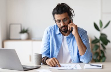 Genç Arap erkek girişimci cep telefonuyla konuşuyor ve modern ofiste kağıtlarla çalışıyor, masamızda oturan yakışıklı bir doğulu adam mali raporları kontrol ediyor ve cep telefonuyla konuşuyor, boş yer var.